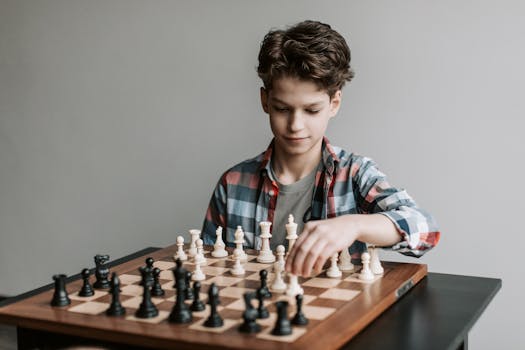 Boy playing chess, deep in thought, sitting at a wooden table indoors.